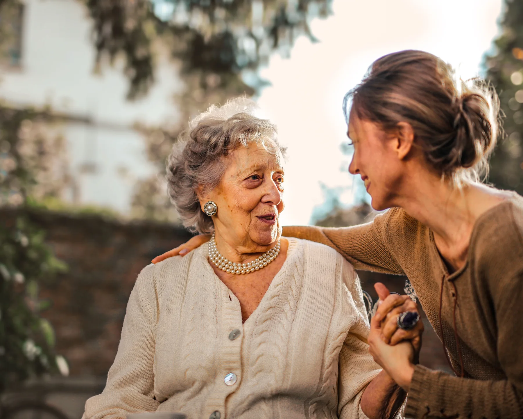 An adult daughter and her senior mother smiling at each other in warm afternoon light in a garden