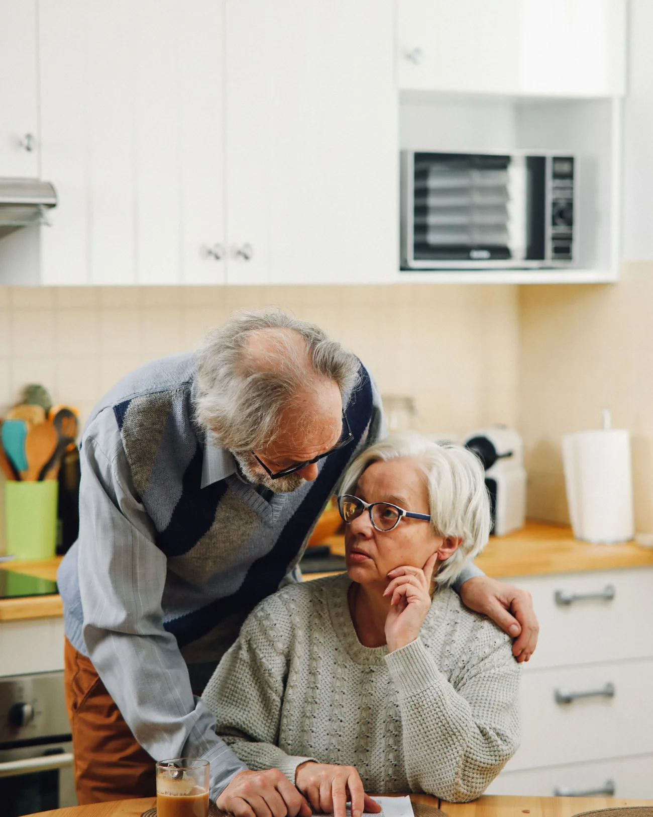An adult son standing beside his seated senior mother in their kitchen, looking at her with concern as they go over paperwork together